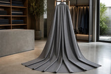 A long, floor-length gray fabric drape or curtain displayed on a hanger in a modern interior showroom with wood shelving, folded garments, and large windows.