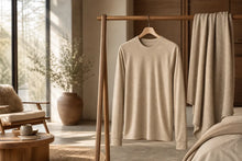 A light beige crewneck sweater hangs on a wooden clothing rack in a softly lit bedroom or lounge setting, with a matching throw blanket draped over the rack and a chair visible in the background.