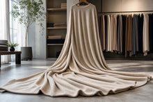 A long, draped cream-colored knit garment or fabric piece hanging on a wooden hanger in a softly lit boutique interior, with shelves, folded textiles, and hanging garments visible in the background.