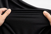 A close-up of a person's hands stretching a piece of black fabric to show its elasticity and texture against a light gray background.