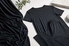 A black two-piece loungewear set consisting of a short-sleeve crewneck top and matching drawstring pants, styled in a flat-lay arrangement with draped black fabric, green leaves, and a stone on a white surface.