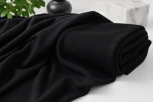 A close-up of a black textured throw blanket draped and folded on a light surface, with a marble block and dark decorative object in the background.
