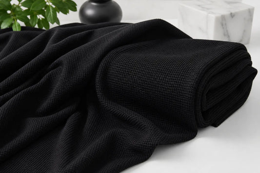 A close-up of a black textured throw blanket draped and folded on a light surface, with a marble block and dark decorative object in the background.