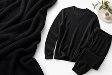 A black long-sleeve crewneck top and matching black pants laid flat on a white surface, styled with a draped black textured blanket and a small ceramic cup with greenery in the corner.