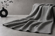 A black and white houndstooth patterned throw blanket draped over a gray concrete surface next to a black ceramic vase with dried stems.