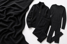 A flat-lay of a black knit clothing set on a white background, featuring a zip-front cardigan, a long-sleeve top, and matching pants, alongside a draped black knit blanket.