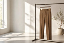 A pair of tailored brown trousers hangs on a minimalist clothing rack in a bright, modern room with natural sunlight casting soft shadows on the wall and floor.