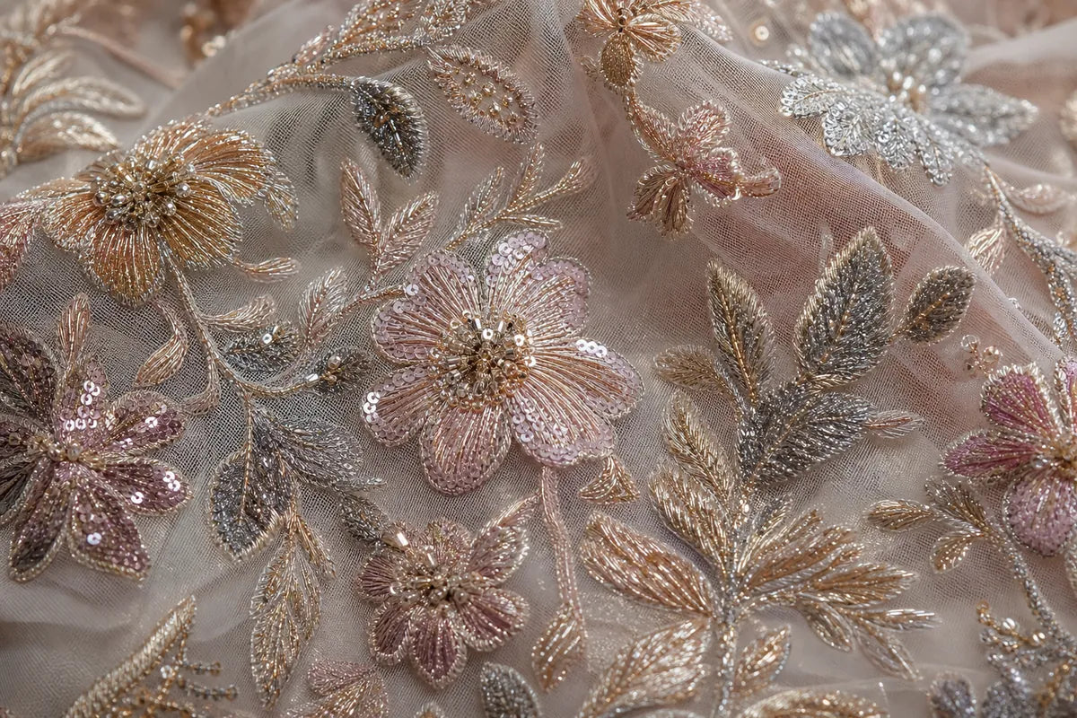 Close-up of a sheer blush pink tulle fabric adorned with intricate floral and leaf embroidery in metallic gold, rose gold, and silver sequins.