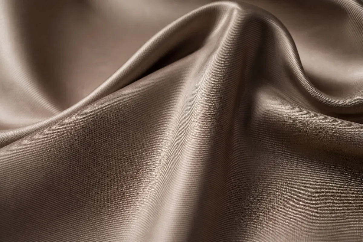 Close-up of draped satin fabric in a warm taupe-brown color, showing soft folds and a subtle sheen.