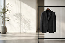 A dark charcoal gray wool-blend blazer jacket hanging on a wooden hanger from a minimalist black clothing rack, set against a bright, modern interior with natural light casting leaf shadows on the wall.