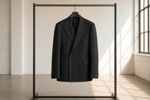 A black double-breasted pinstripe blazer hanging on a wooden hanger from a minimalist black clothing rack, set against a bright, sunlit studio background.