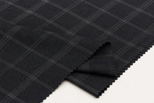A close-up of dark charcoal gray fabric with a fine white windowpane plaid pattern, folded over on a white surface.