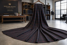 A long, dark plaid tailcoat coat with a matching long tail is displayed on a wooden hanger in a modern, stylishly decorated tailoring studio. The coat features a dark navy and black base with red and subtle gold lines in the plaid pattern.