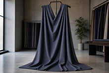 A dark slate blue draped fabric, likely a velvet or velour upholstery material, hanging from a wooden hanger in a minimalist, modern studio or showroom with concrete floors and walls.