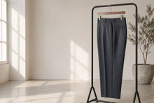 Dark gray tailored trousers hanging on a black clothing rack in a bright, minimalist room with natural window light and a potted plant in the background.