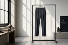 Dark charcoal gray pinstriped trousers hanging on a minimalist black clothing rack in a modern, sunlit room with a window and decorative vases.
