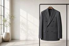 A dark gray double-breasted pinstripe suit jacket hanging on a wooden hanger from a black clothing rack in a bright, modern room with natural light.