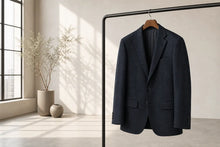A dark navy blue textured blazer jacket hanging on a wooden hanger from a black clothing rack, set in a minimalist room with a window and dried plants.