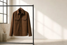 A brown long-sleeve button-up shirt jacket with two chest flap pockets, hanging on a black clothing rack in a bright, minimalist room with window light casting shadows on the wall.
