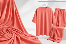A coral pink matching lounge set consisting of a short-sleeve crewneck t-shirt on a hanger and matching drawstring sweatpants, displayed alongside a large draped fabric swatch of the same material on a white background.
