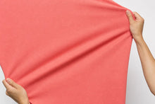 A coral pink fabric sheet being held stretched by two hands against a plain light gray background.