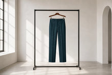 A pair of dark teal trousers hanging on a wooden hanger from a minimalist black clothing rack, set in a bright, modern room with natural light coming from a window.