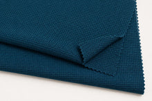 A close-up of folded dark teal blue textured fabric with a waffle or grid-like weave and scalloped edges, shown on a plain white background.