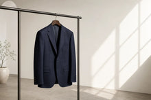 A navy blue suit jacket hangs on a wooden hanger from a minimalist black clothing rack in a bright, modern room with warm sunlight casting diagonal shadows on the wall.