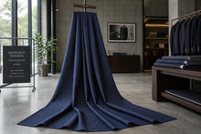 A large draped piece of dark navy blue fabric with fine vertical pinstripes, displayed on a mannequin stand inside a modern clothing store or showroom. The background features other garments on racks, a display table, and a sign that reads 'DENIM BLUE PINSTRIKE'.