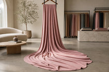 A dusty pink knit blanket or throw draped over a hanger in a modern, minimalist boutique interior, with other fabric swatches and furniture visible in the background.