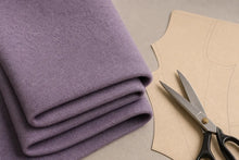 A folded piece of textured lavender-purple fabric next to a beige paper pattern and silver scissors on a neutral gray surface.