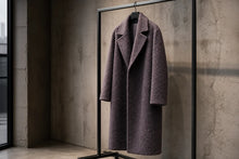 A textured, mid-length charcoal gray coat with wide lapels hangs on a black metal clothing rack against a concrete wall.