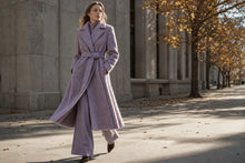 A woman walking outdoors in a long lavender-purple belted coat and matching wide-leg trousers, with a stone building and trees in the background.