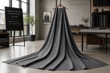 A dark gray structured wool coat draped over a wooden hanger in a modern, stylish interior studio setting. The coat features a dramatic, long train and is displayed against a background with a chalkboard detailing fabric specifications, a framed portrait, and shelves.