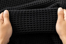 A close-up of a black waffle-weave fabric being held up by two hands to show its texture and flexibility.
