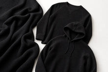 Three black knit garments laid flat on a white background: a draped textured knit fabric piece on the left, a long-sleeve crewneck top in the back, and a hooded pullover in the front.