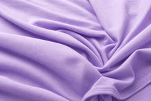 A close-up of lavender purple fabric with soft folds and a stitched hem visible in the lower right corner.
