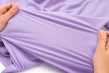 A close-up of lavender purple fabric being held and stretched by two hands to show its texture and elasticity.