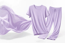 A lavender purple two-piece pajama set consisting of a short-sleeve crewneck t-shirt and matching drawstring pants, shown floating against a clean white background with a matching fabric sheet draped on the left.