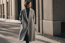 A woman wearing a long, light gray wool coat over a black turtleneck walks along a city sidewalk beside a modern concrete building.