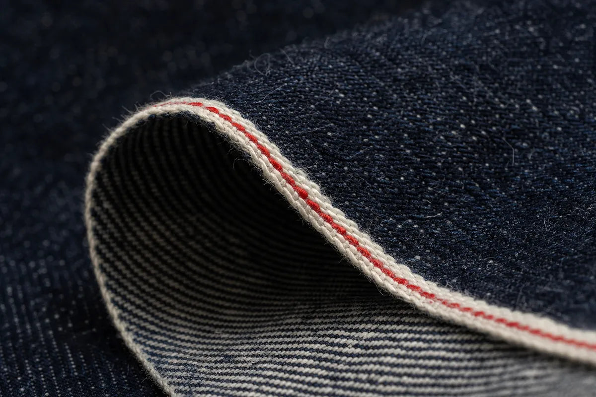 Close-up of dark indigo denim fabric rolled over, showing the textured weave and a contrasting red-and-white whipstitch edge.