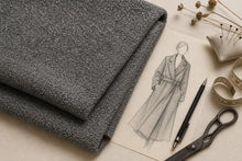 Folded gray textured fabric displayed on a beige surface next to a pencil sketch of a trench coat, with a pen, measuring tape, pins, and dried flowers.