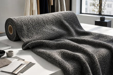 A dark charcoal gray textured throw blanket draped over a white table, with a matching rolled fabric sample beside it in a bright, modern room setting.