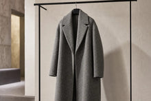 A long gray wool-blend coat hanging on a black clothing rack against a textured off-white wall, with a softly blurred interior space visible on the left.