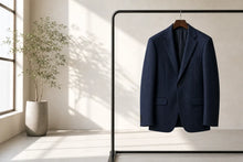 A navy blue wool blazer jacket hanging on a wooden hanger from a minimalist black clothing rack, set in a bright, modern room with natural sunlight casting soft shadows on a beige wall.