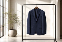 A navy blue tailored blazer hangs on a wooden hanger from a black clothing rack in a bright, modern interior with sunlight streaming through a window and a potted plant nearby.