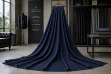 A long navy blue plaid fabric drape hangs from a wooden hanger in a styled interior room with clothing racks, a sign reading 'LINEN CLOSET', and a leather chair visible in the background.