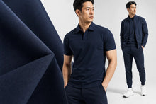 A man modeling a dark navy short-sleeve polo shirt and matching navy pants against a light gray studio background, with a large folded navy fabric panel on the left and a smaller rear view of the same outfit on the right.