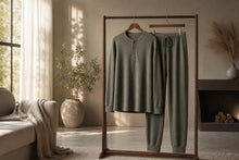 A two-piece lounge set in a muted sage green color, consisting of a long-sleeve henley shirt and matching drawstring pants, displayed on a wooden clothing rack in a cozy, sunlit living room.