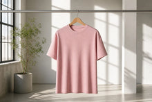 A light pink short-sleeve crewneck t-shirt hanging on a wooden hanger from a metal clothing rack, set in a minimalist room with a potted plant and window shadows.